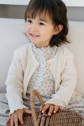 Baby wearing Avauma off white cardigan and beige ribbed pants, sitting on a wooden chair